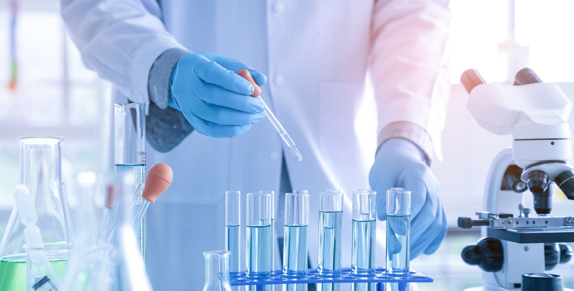 Scientist in lab coat and gloves, using a pipette to add liquid to a test tube.