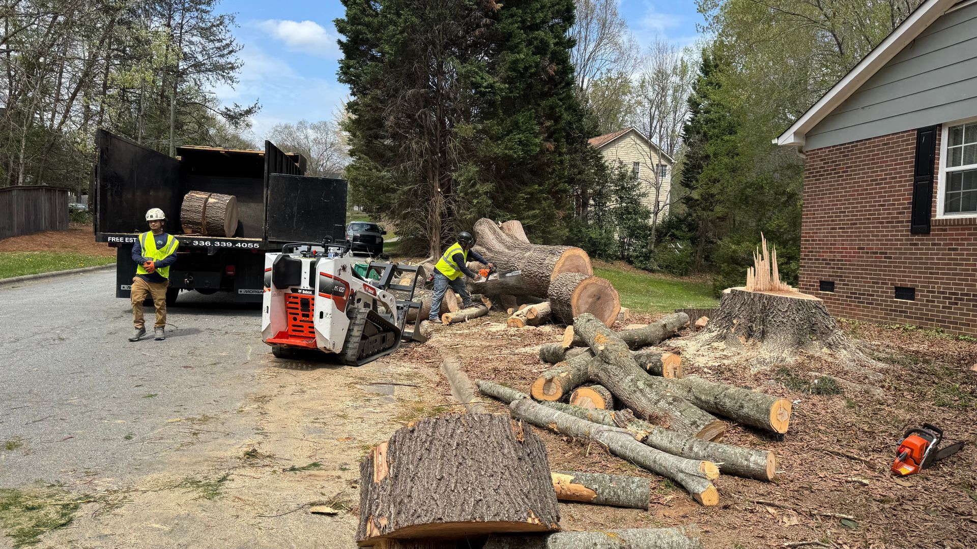 Workers clearing fallen tree trunks along a driveway beside a house and truck, with a chainsaw and wood chips scattered.