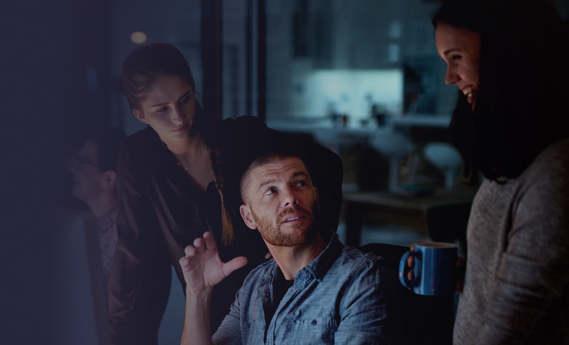Three people collaborating in a dimly lit room, two women looking at a man who gestures animatedly.