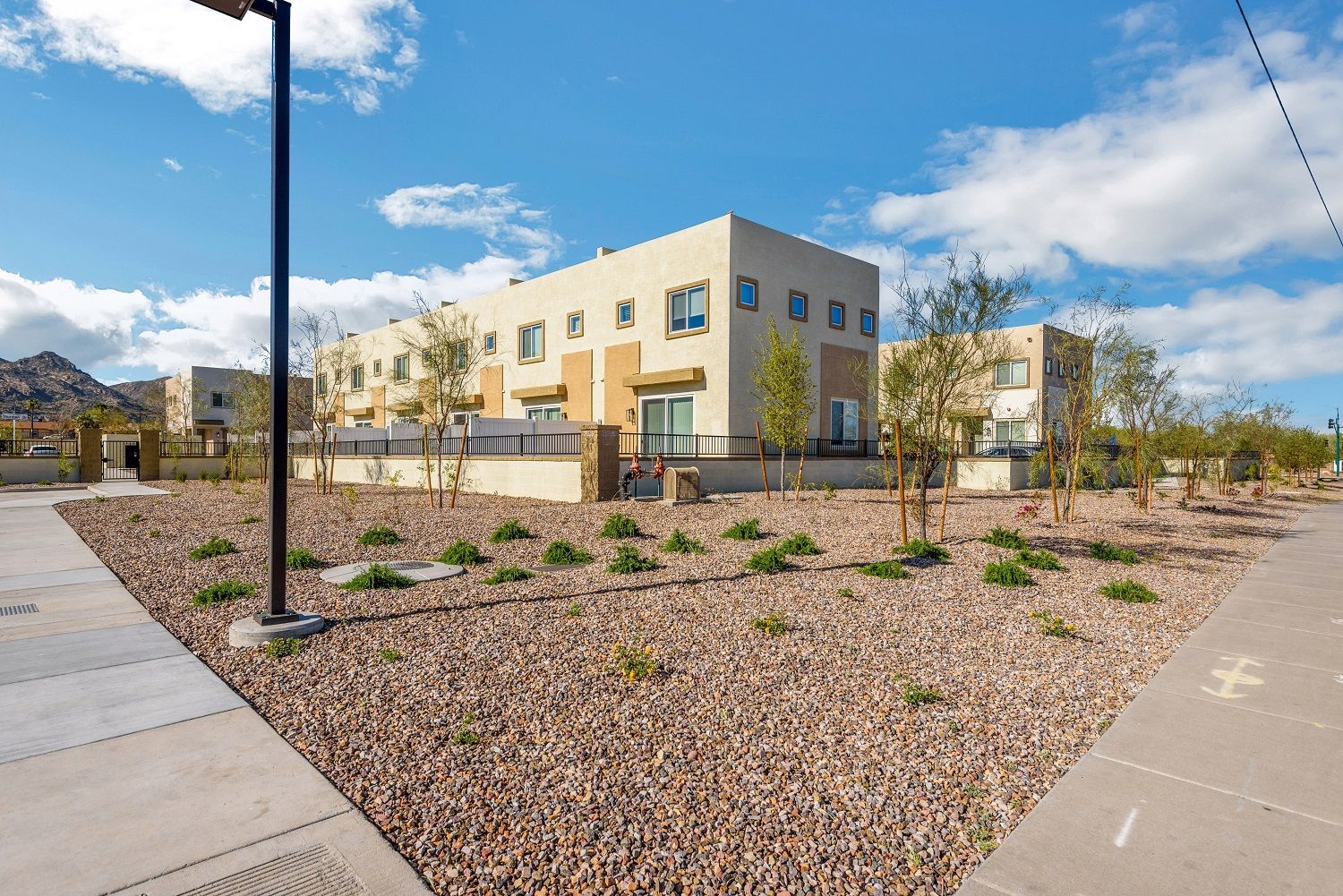 Modern beige apartment buildings beside a landscaped gravel courtyard and sidewalks under a blue sky