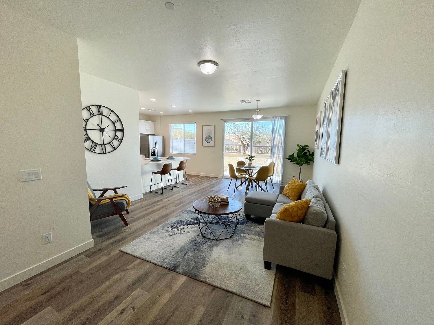 Bright living room with gray sofa, rug, coffee table, wall clock, and a view of trees through large windows