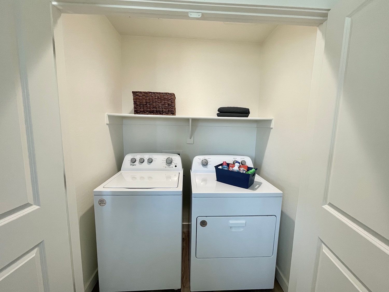 Laundry closet with a white washer and dryer, shelf above, and folded items on top.