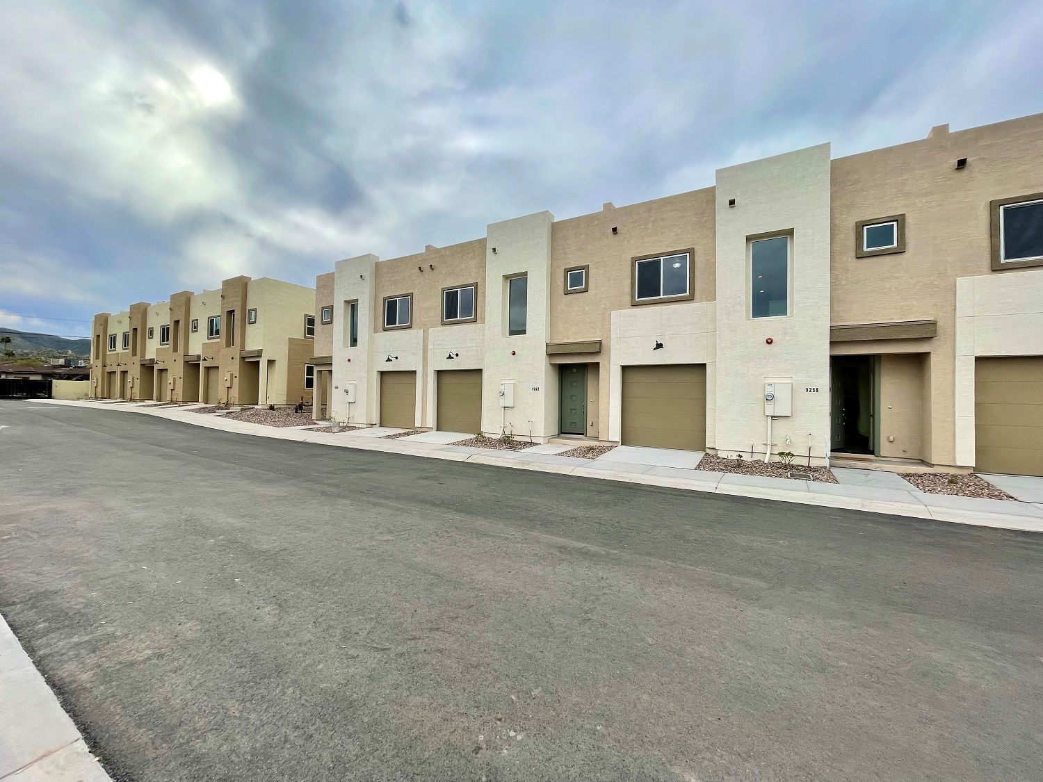 Row of beige townhouses with garages along a wide empty street under a cloudy sky
