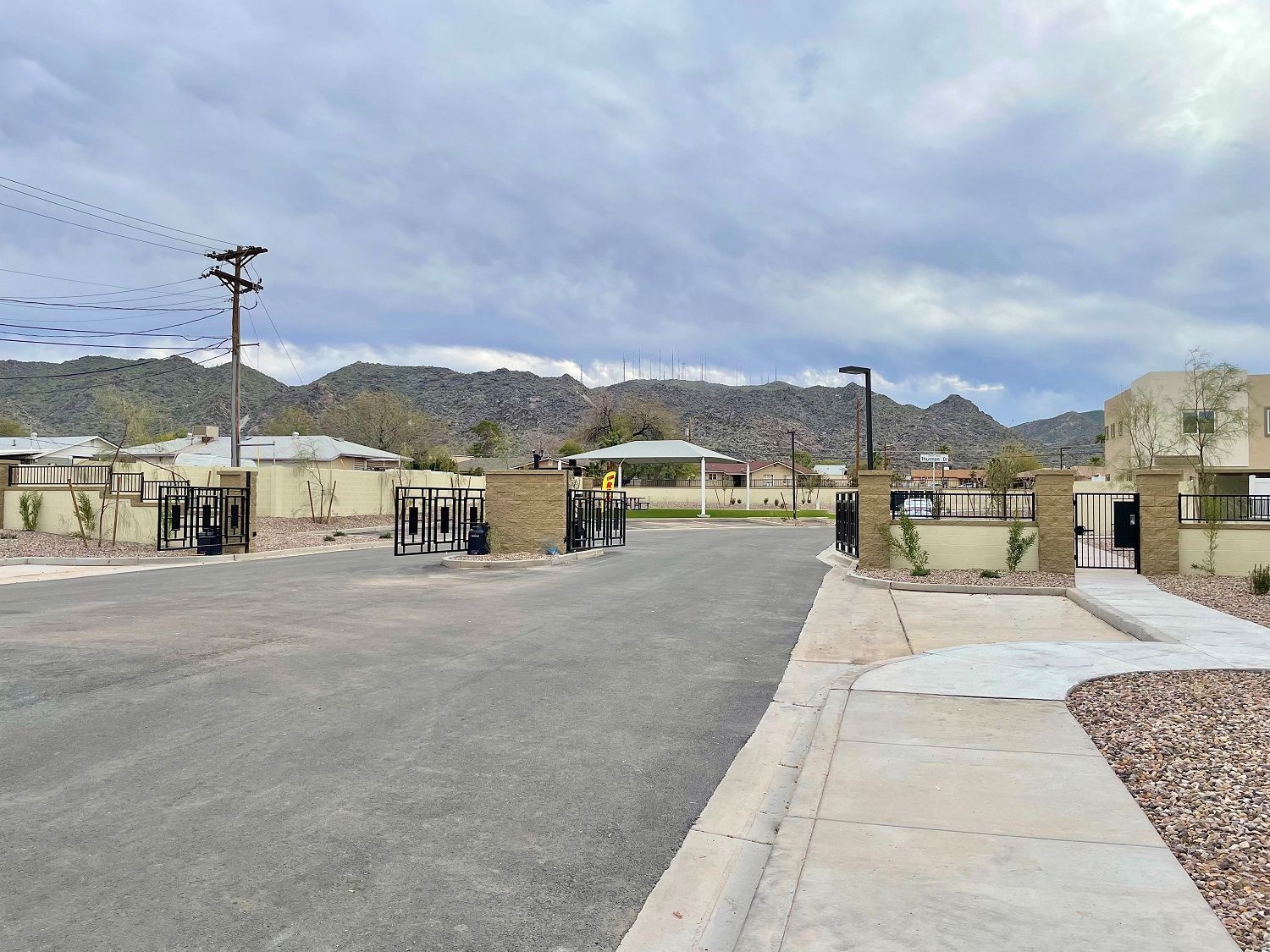 Gated entrance to a residential community with paved road, stone pillars, and mountains under cloudy sky