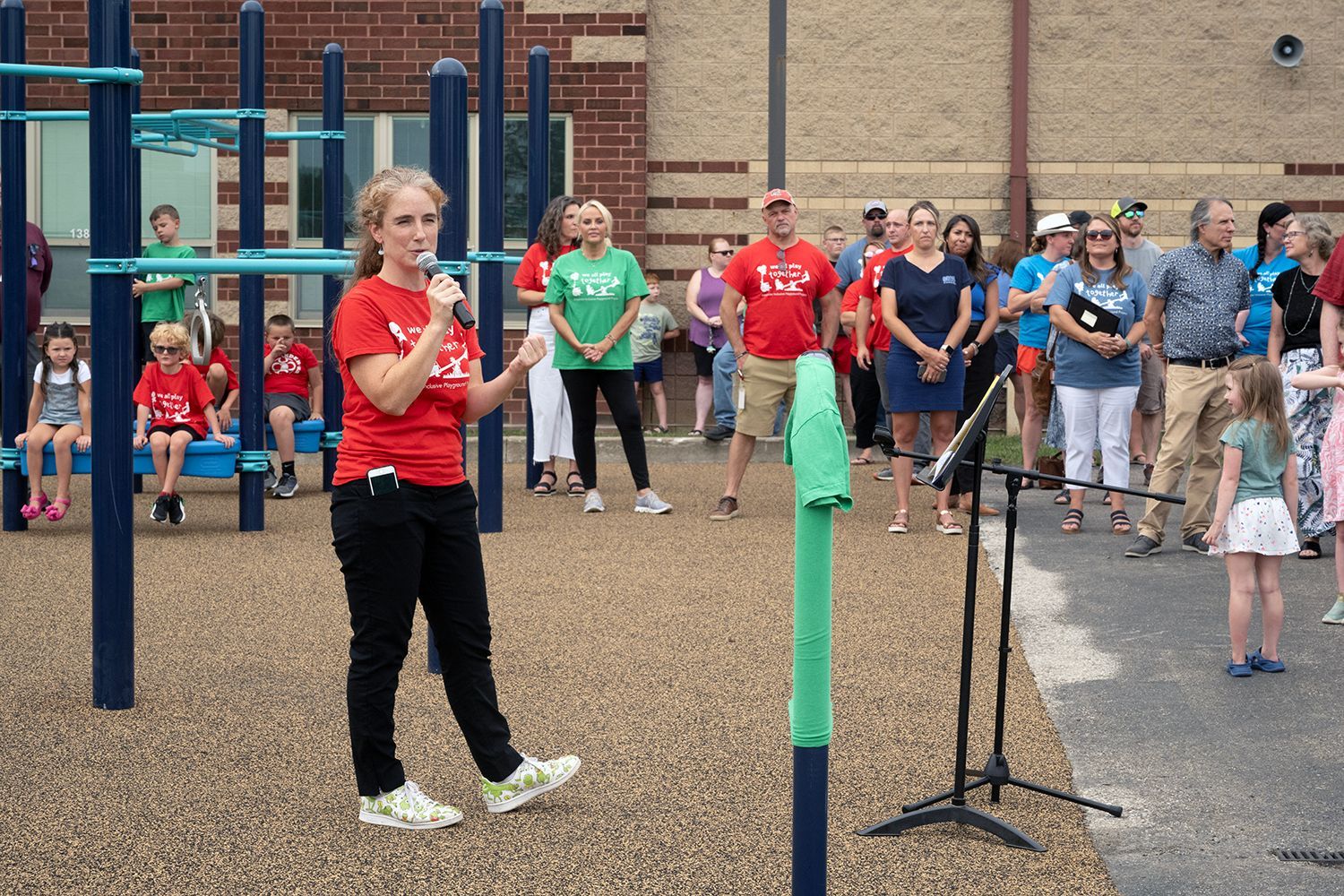 Woman in red shirt speaks into a microphone at a playground opening, crowd watches.