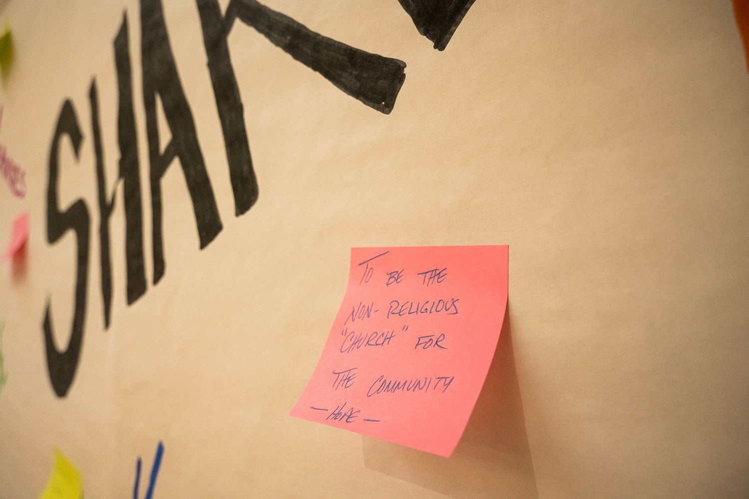 Pink sticky note on a wall, with handwritten text and part of the word 