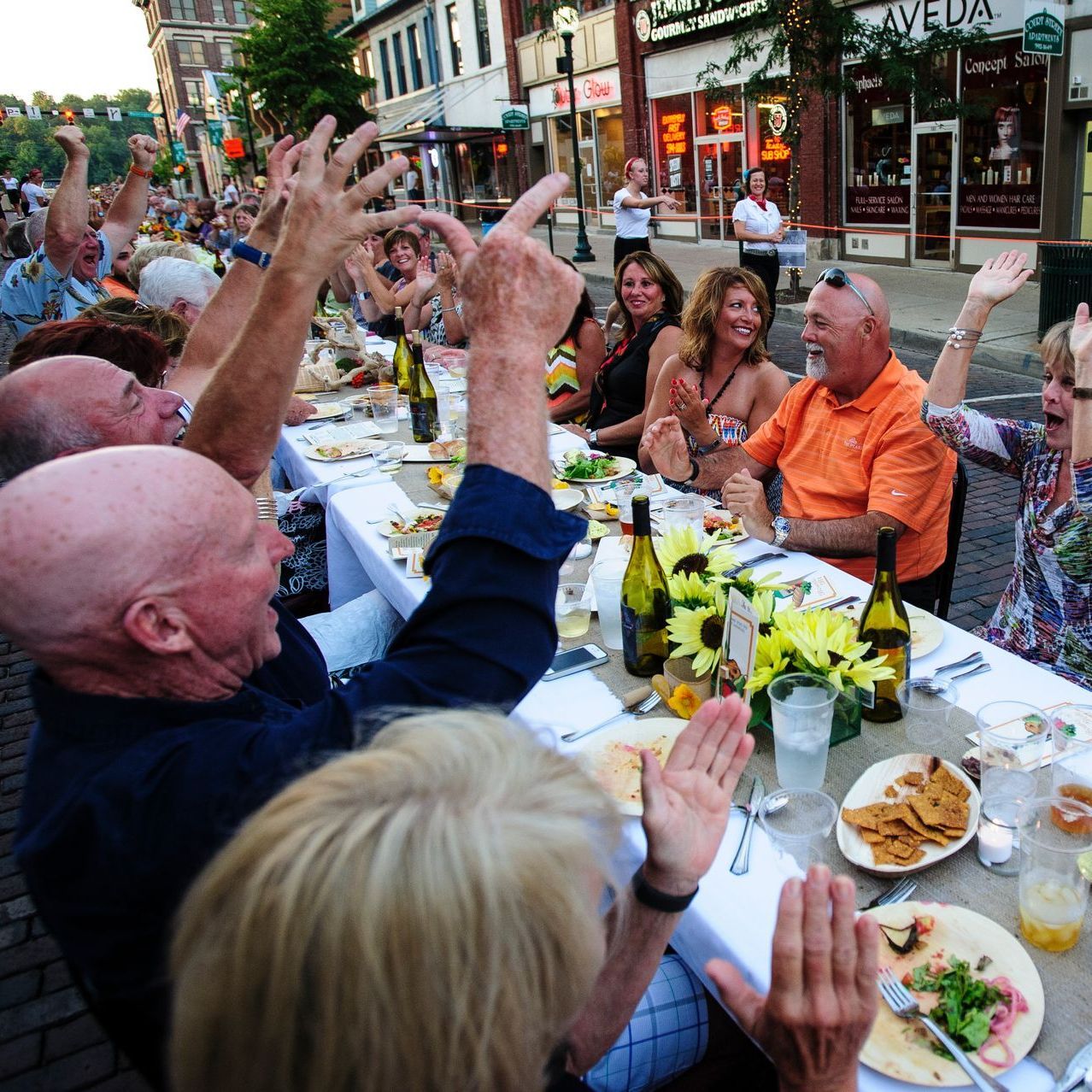 People cheering at a long table set for a meal outdoors on a city street. Bright light, food, drinks. Bounty on the Bricks, Athens County, OH