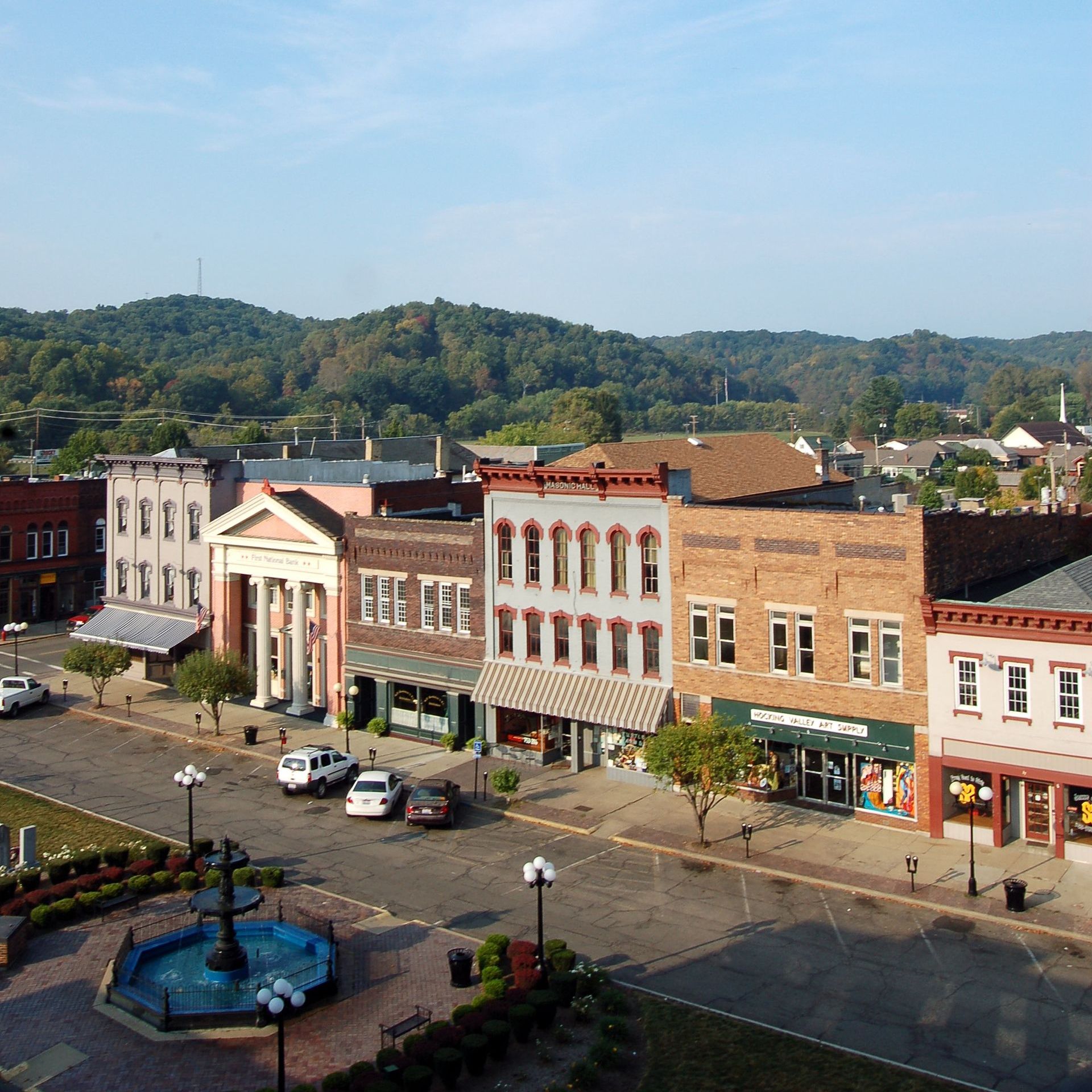 Nelsonville City square with historic buildings, cars, fountain, and green hills in the background. Athens County, OH