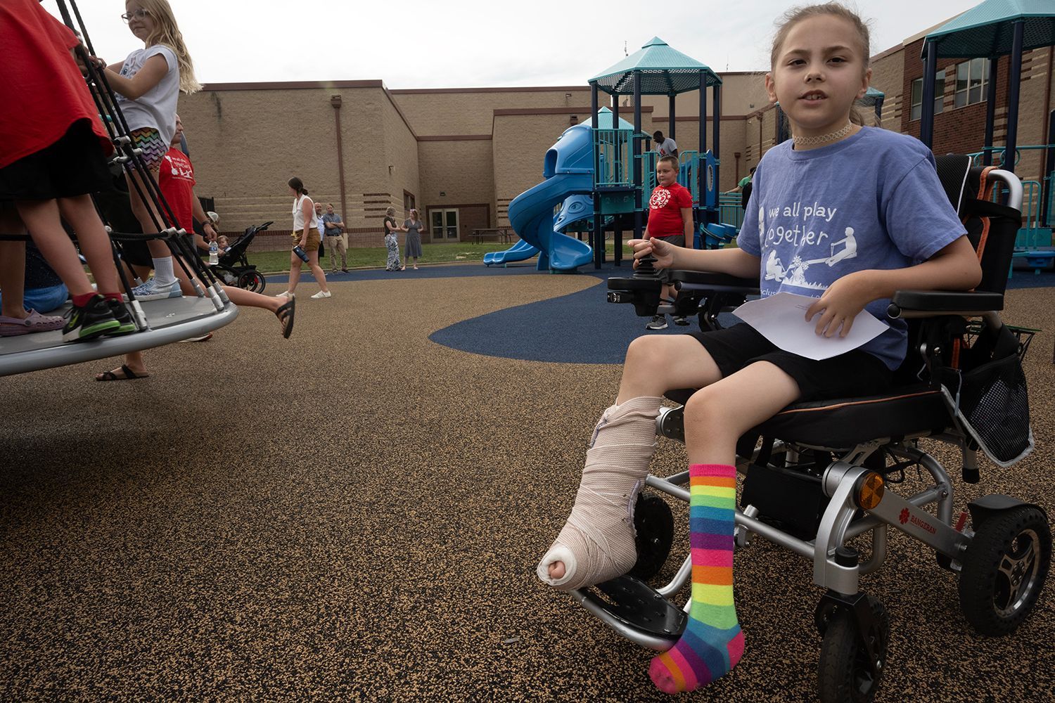 Girl in wheelchair with leg casts at a playground, wearing rainbow socks, looking at the camera.
