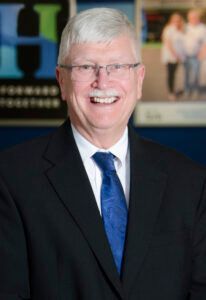 Smiling man in suit, glasses, and blue tie. Background has blue, green, and white elements.