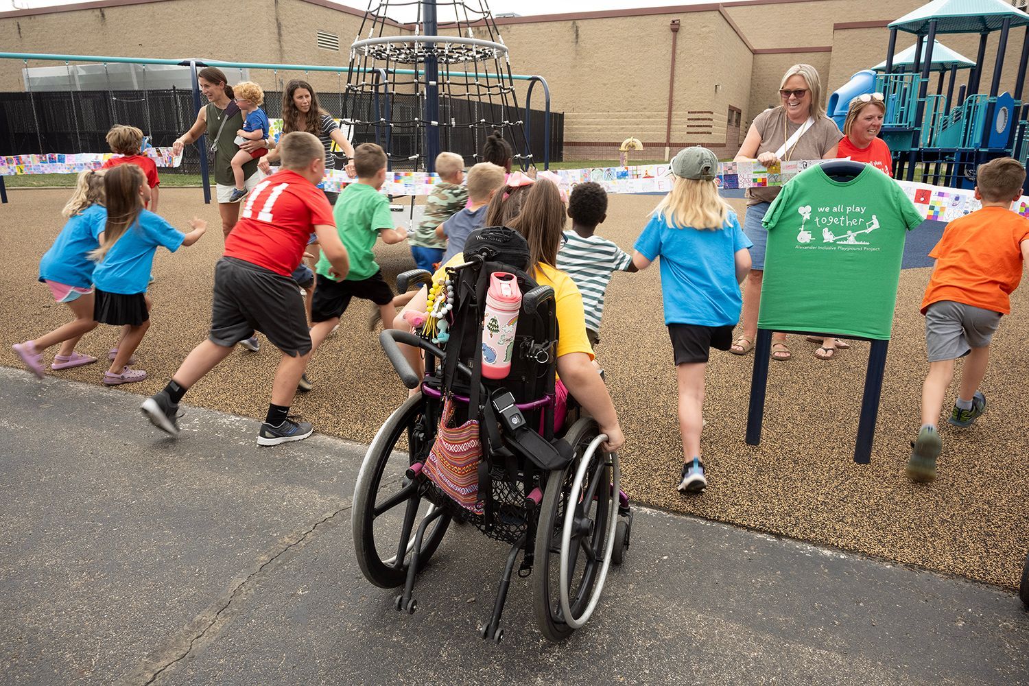 Children playing on a playground, one in a wheelchair. Others run and interact with adults.