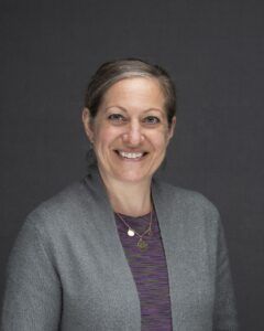 Woman with gray hair and a warm smile, wearing a gray sweater and purple shirt, against a gray background.