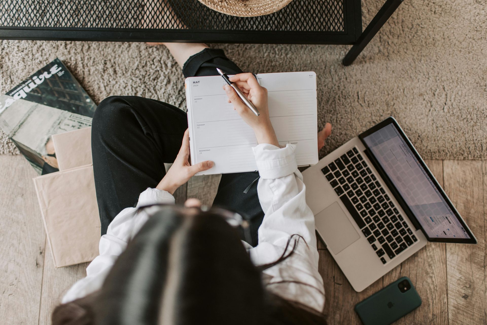 Woman sitting on floor, writing in calendar with pen, laptop and phone nearby.