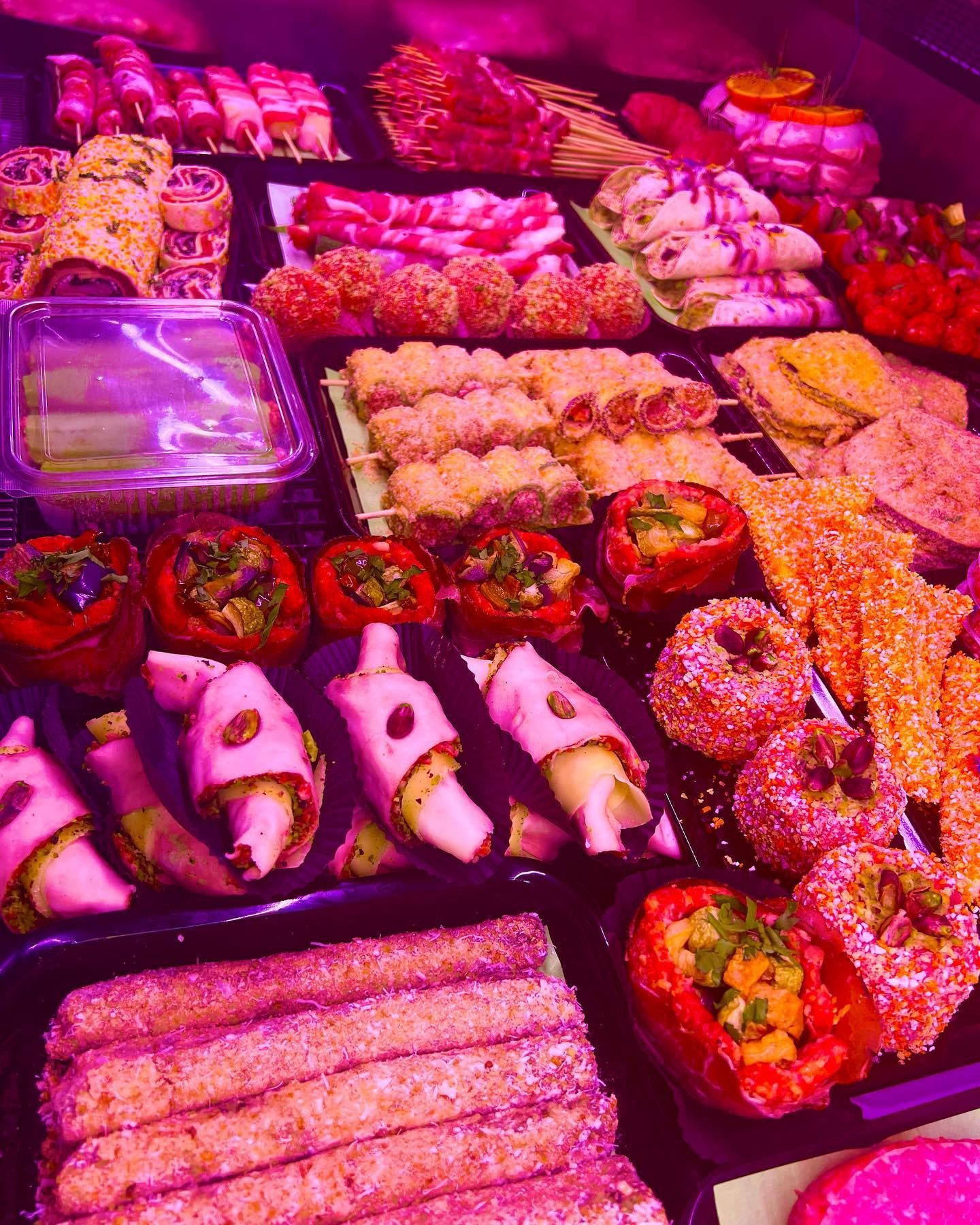 Sushi assortiti e involtini al forno disposti su vassoi sotto luci rosa in un'esposizione di prodotti alimentari.