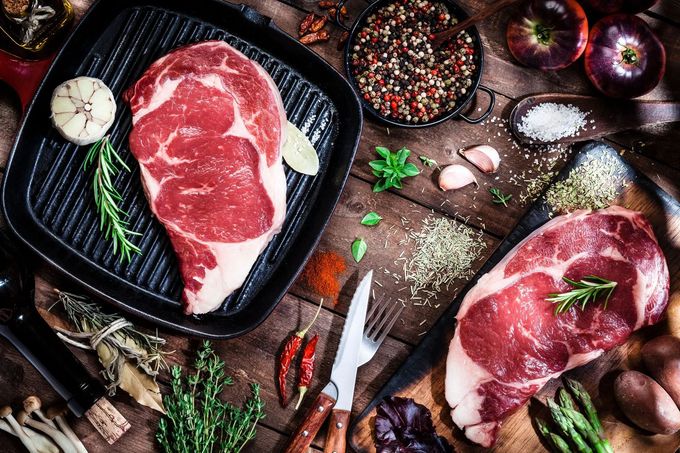 Due bistecche di costata crude su un tavolo rustico con erbe aromatiche, spezie, aglio e un termometro per carne.
