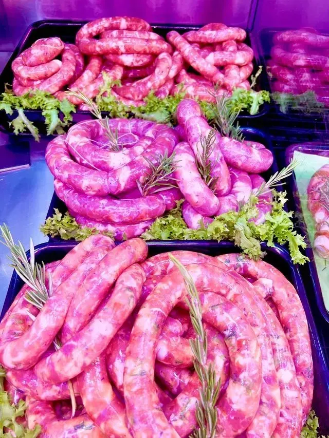 Salsicce viola arrotolate in vassoi in un'esposizione di mercato, guarnite con erbe aromatiche verdi.