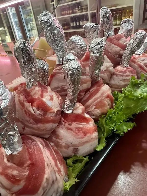 Piatto di spiedini avvolti in pancetta cruda e coperti con un foglio di alluminio, disposti su un letto di lattuga in una vetrina.
