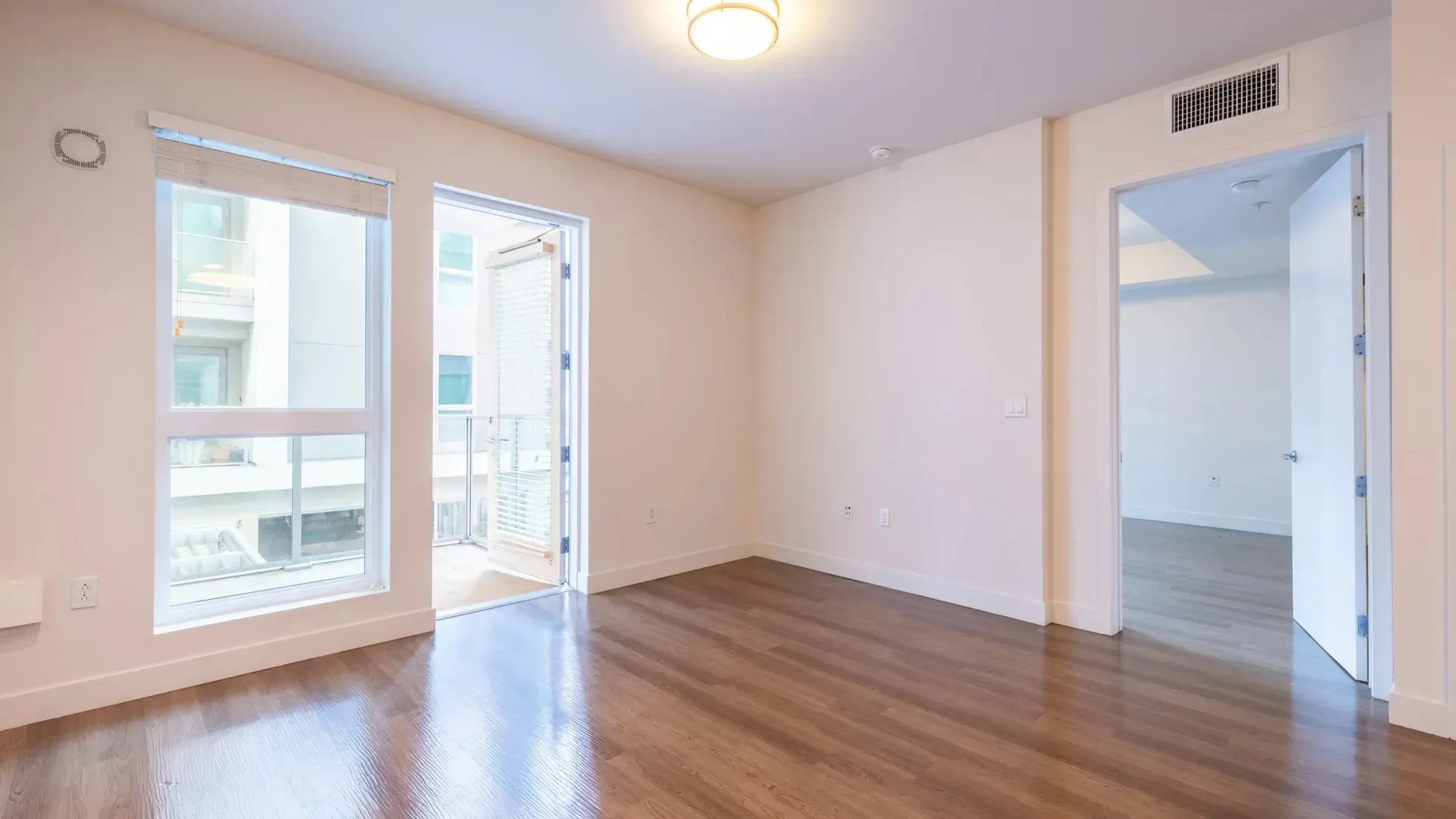 Interior view of a vacant apartment living room with hardwood floors, large windows, and a balcony door.