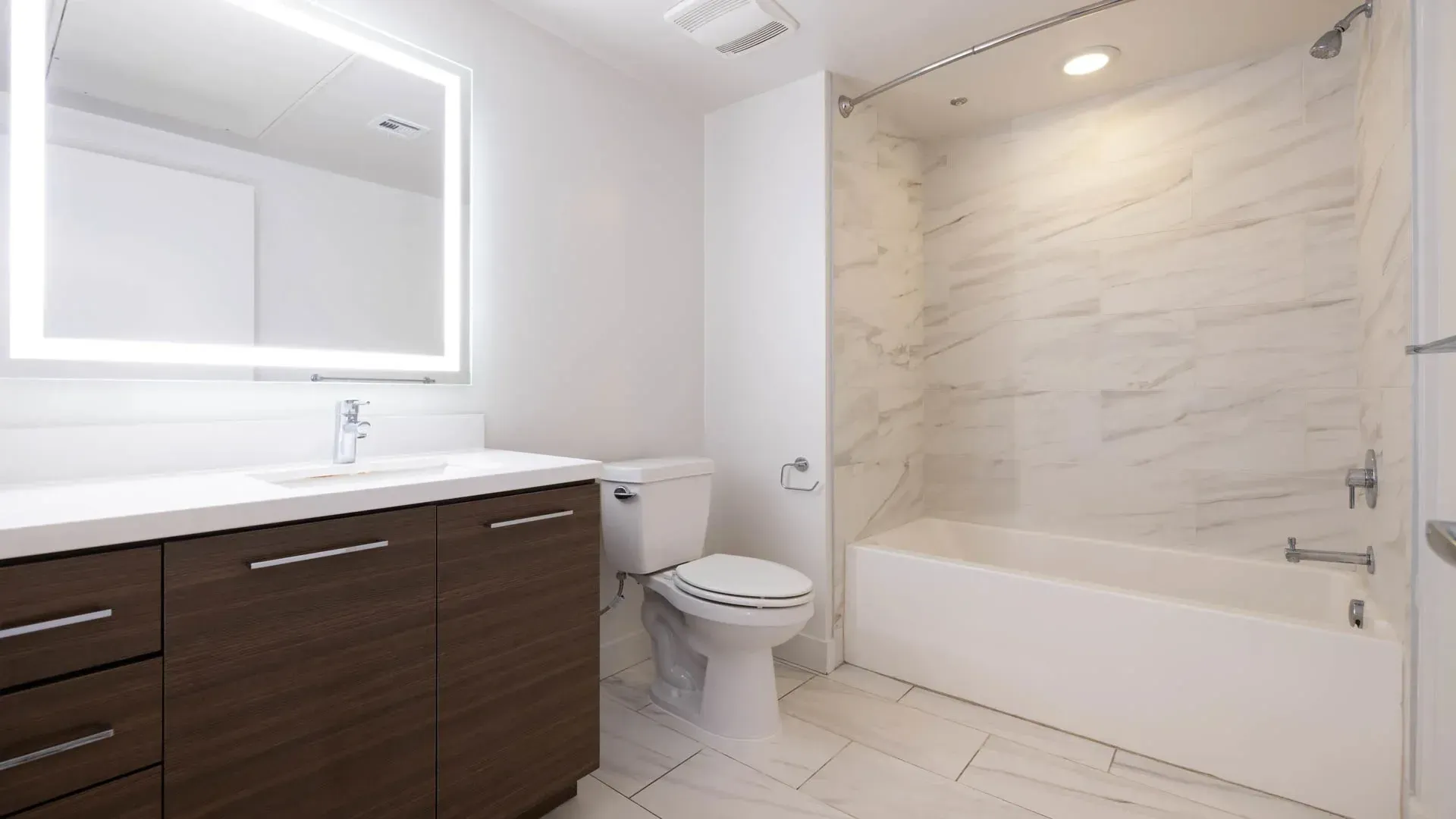 Modern bathroom with a vanity, illuminated mirror, toilet, and a bathtub with marble tile shower surround.
