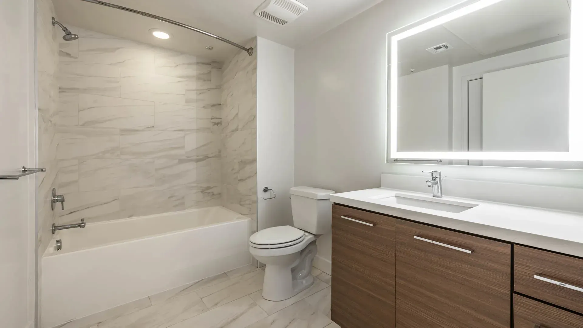 Modern bathroom with a marble tile shower, bathtub, toilet, and vanity with a lit mirror.