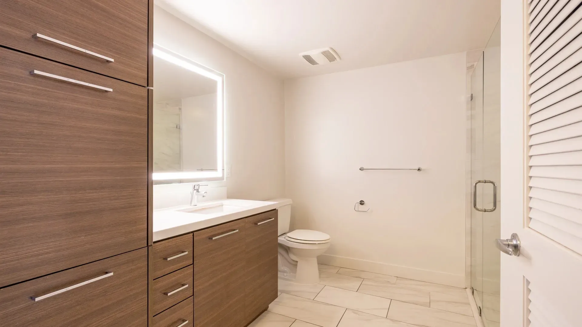 Modern bathroom with wood vanity, illuminated mirror, toilet, and glass-enclosed shower.
