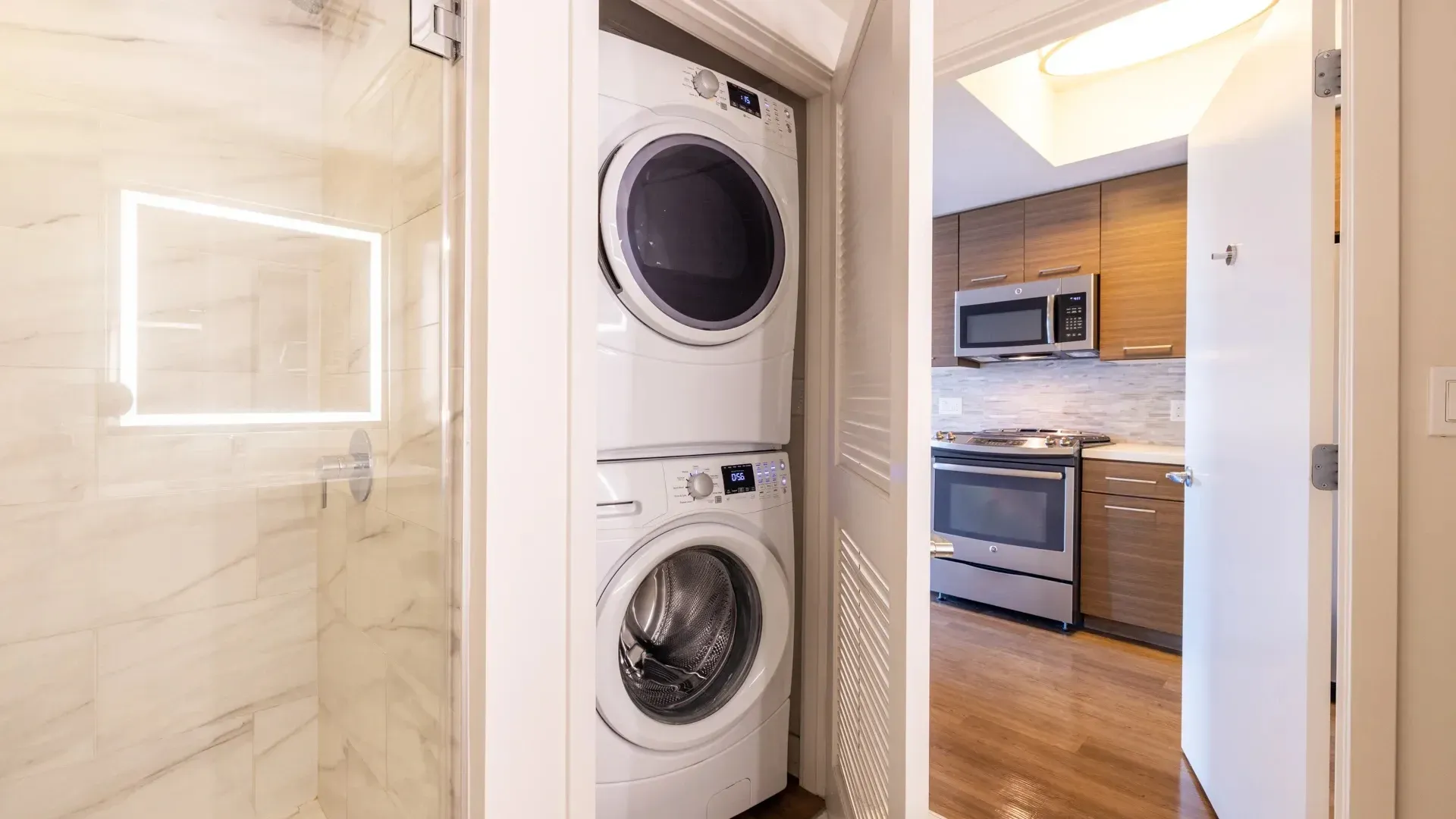Stacked washer and dryer next to a marble shower and a kitchen with stainless steel appliances.