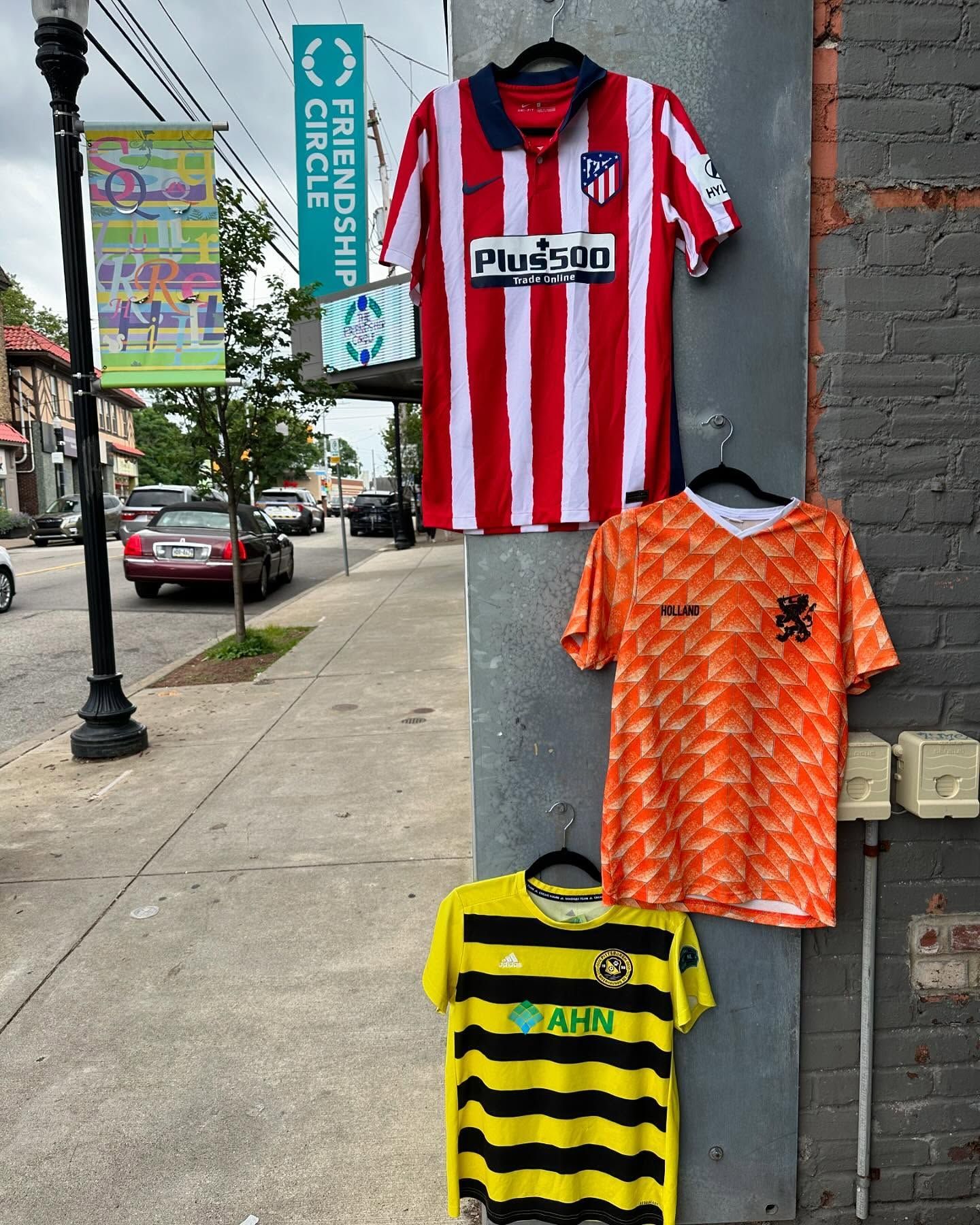 Soccer jerseys hanging outside a shop on a sidewalk, including red-white stripes, orange, and yellow-black.