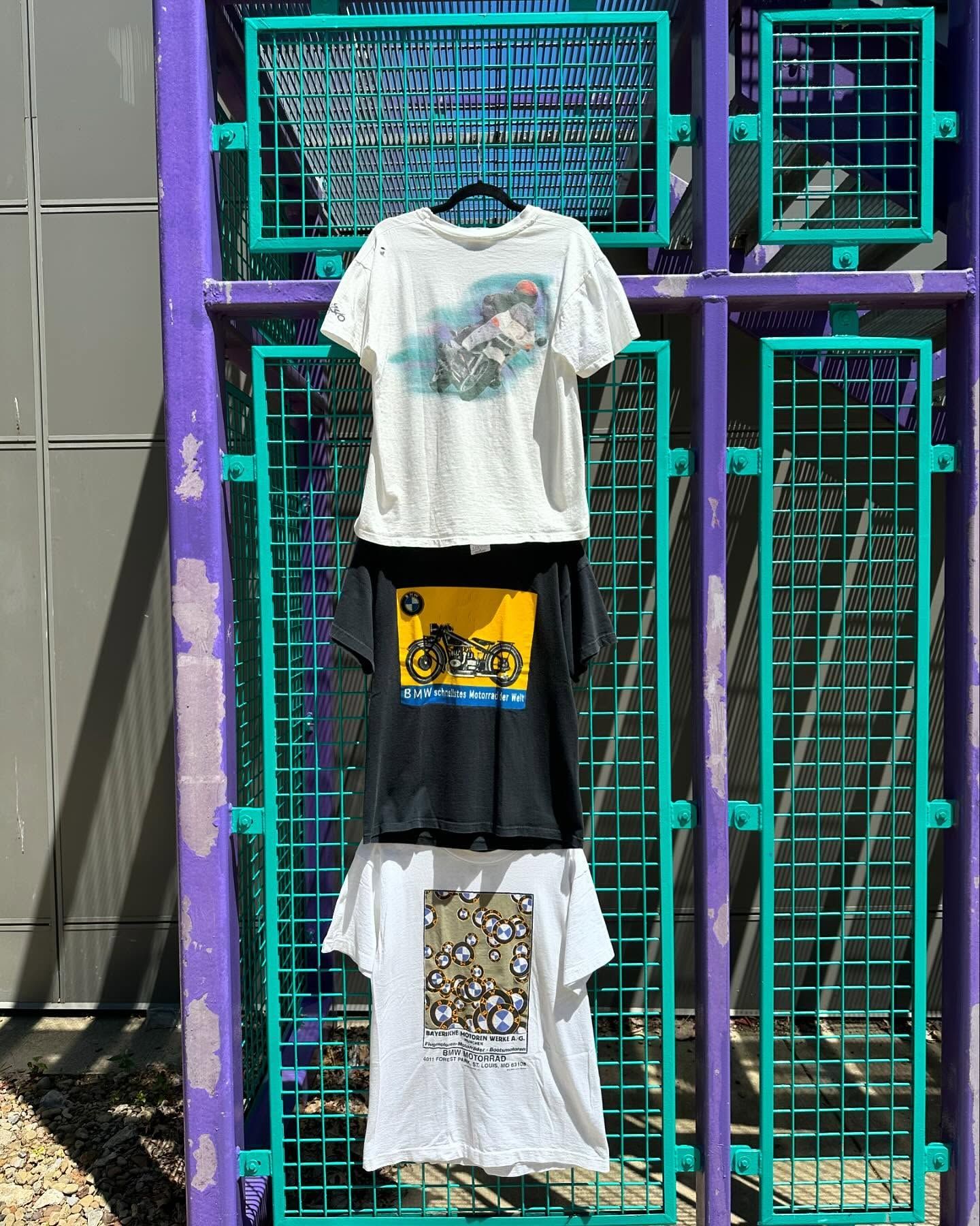 Three graphic T-shirts hanging on a purple storefront gate with teal wire shelves