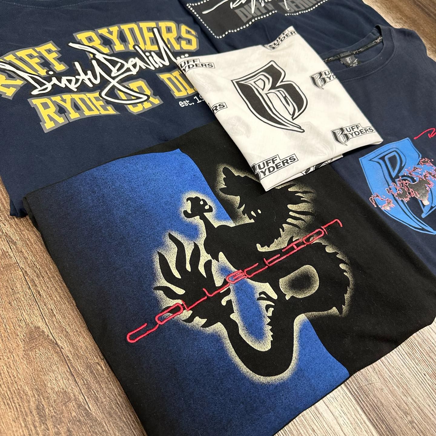 Assorted folded graphic T-shirts in black, blue, white, and tan with bold printed logos and designs