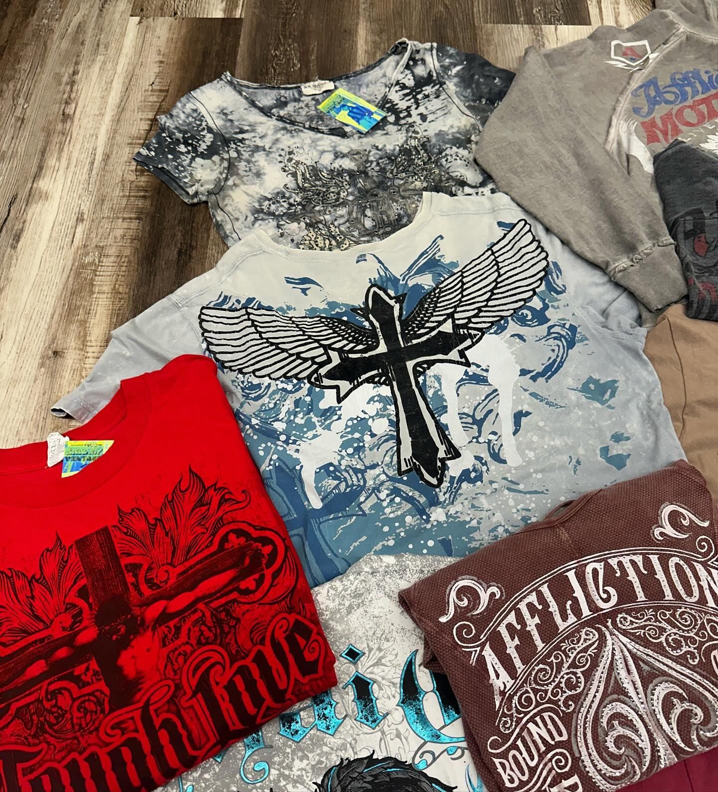 Assorted graphic T-shirts laid out on a wooden floor, including red, gray, brown, and black designs.