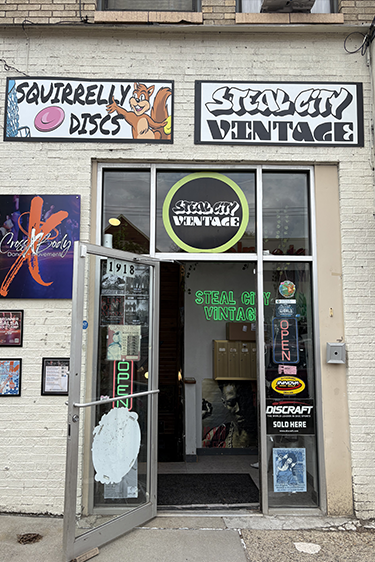 Vintage clothing shop storefront with open door, graffiti-style signs, and colorful window displays