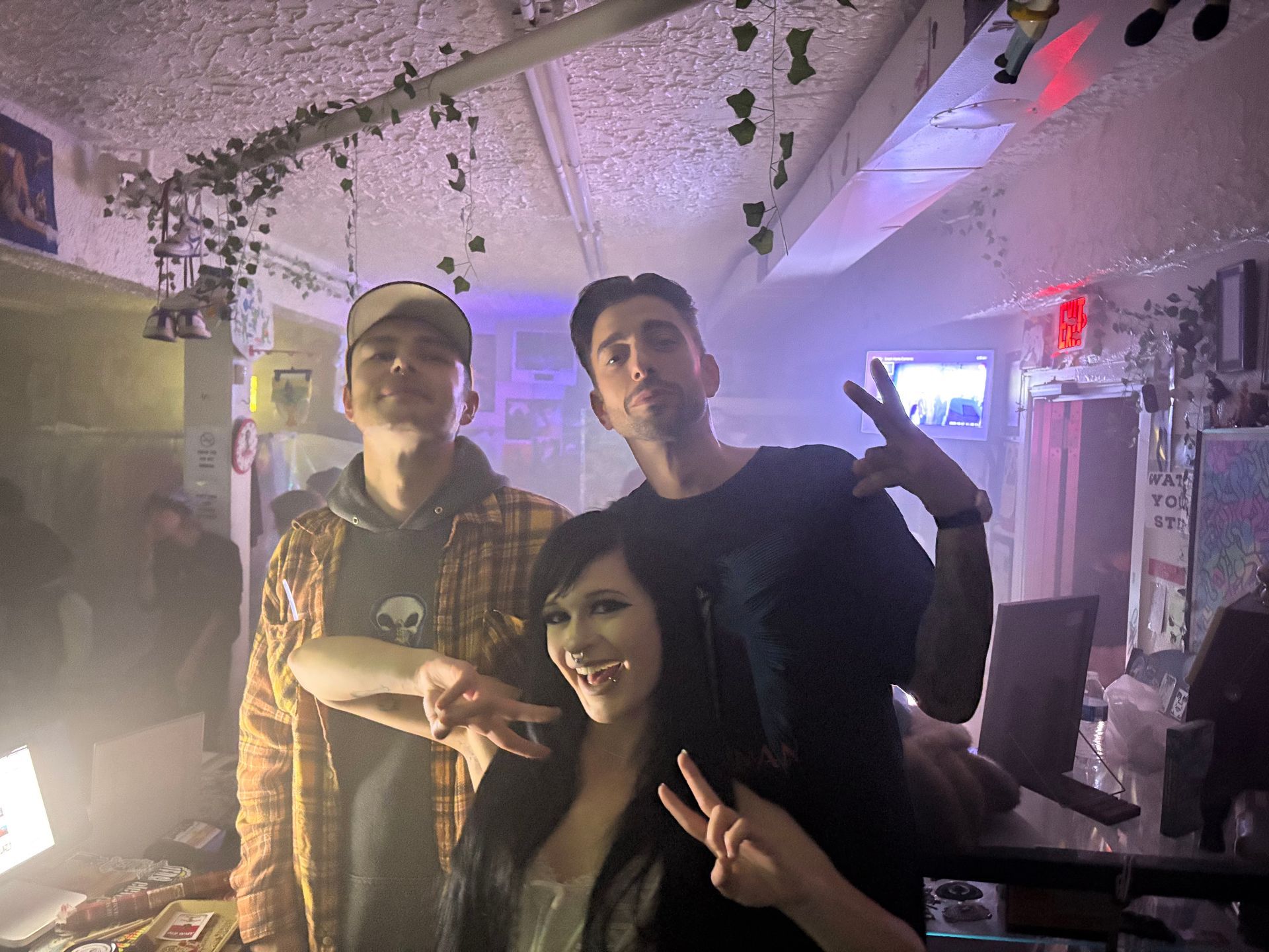 Three people posing indoors under colorful lights, smiling and making peace signs.