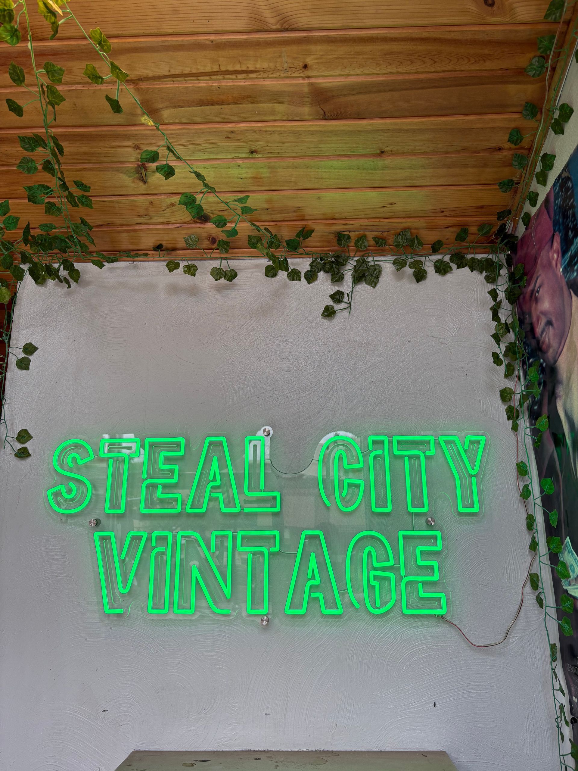 Neon green “STEAL CITY VINTAGE” sign on a gray wall with hanging vines and a wooden ceiling