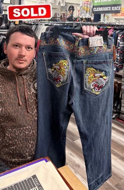 Man holding up a pair of blue jeans with colorful embroidered back pockets in a clothing shop