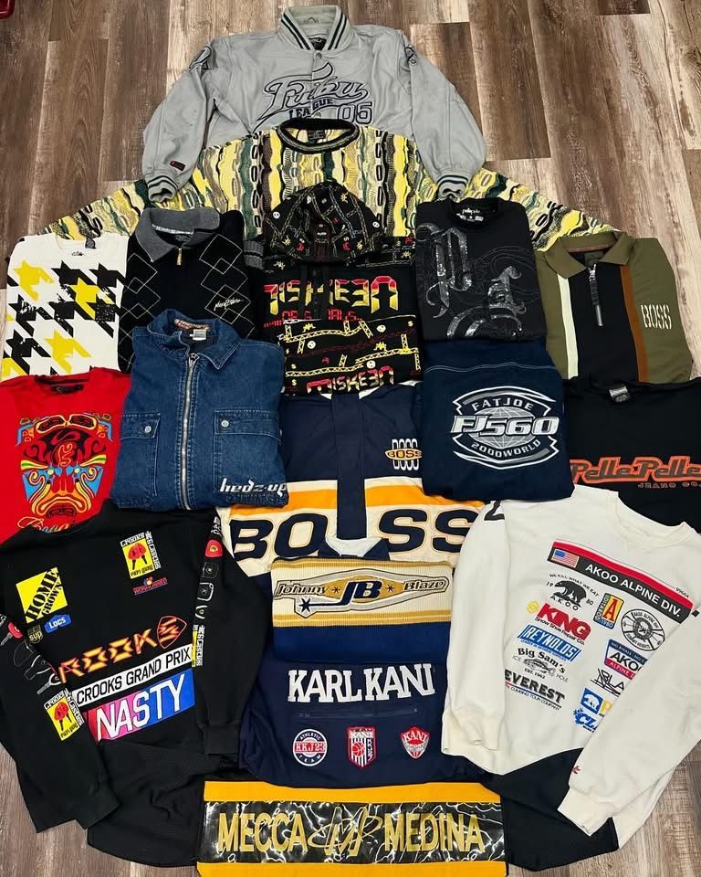 Assorted folded streetwear hoodies, jeans, and graphic long-sleeve shirts laid out on a wooden floor.
