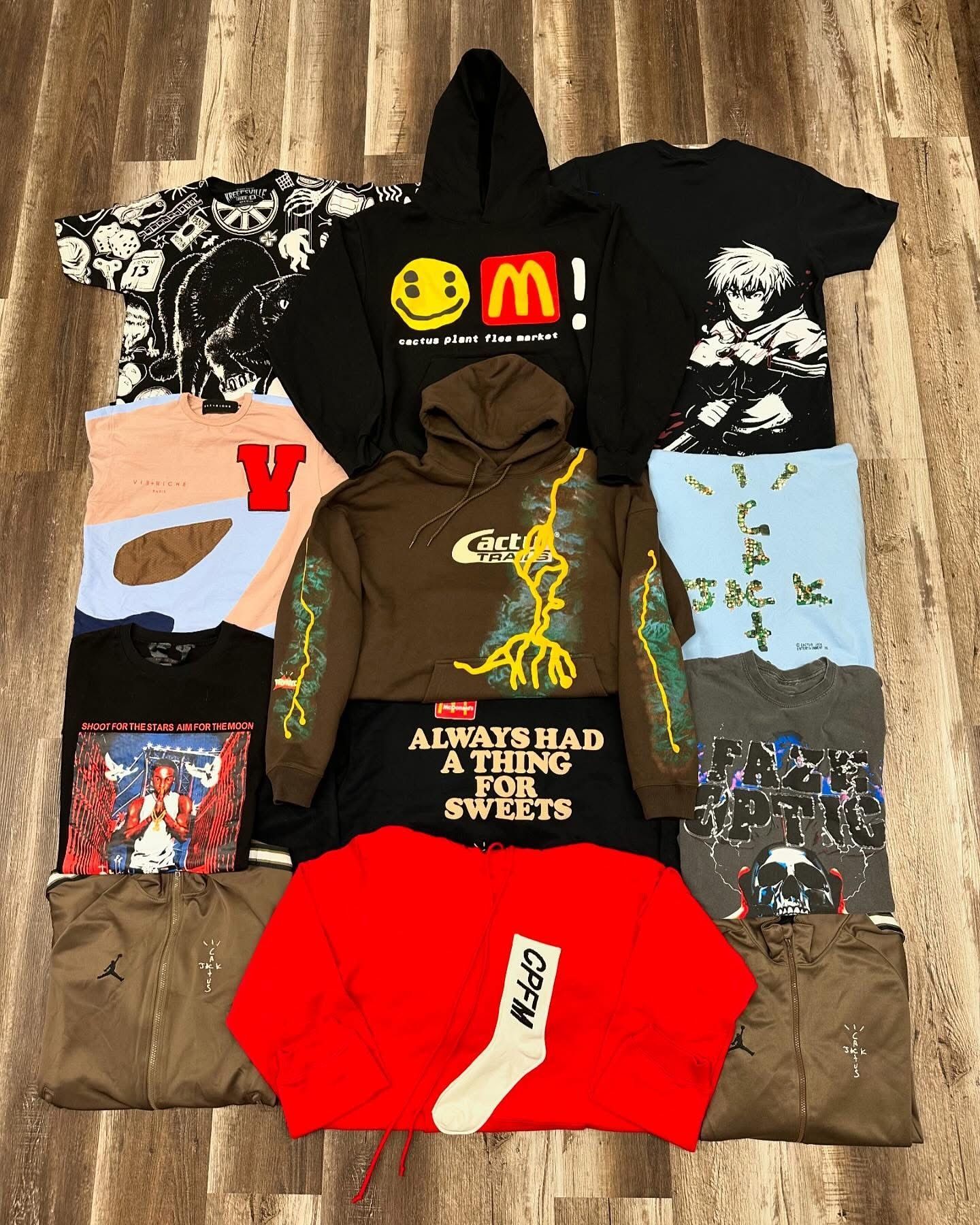 Assorted graphic hoodies and tees laid out on a wooden floor, featuring black, red, brown, and blue designs.