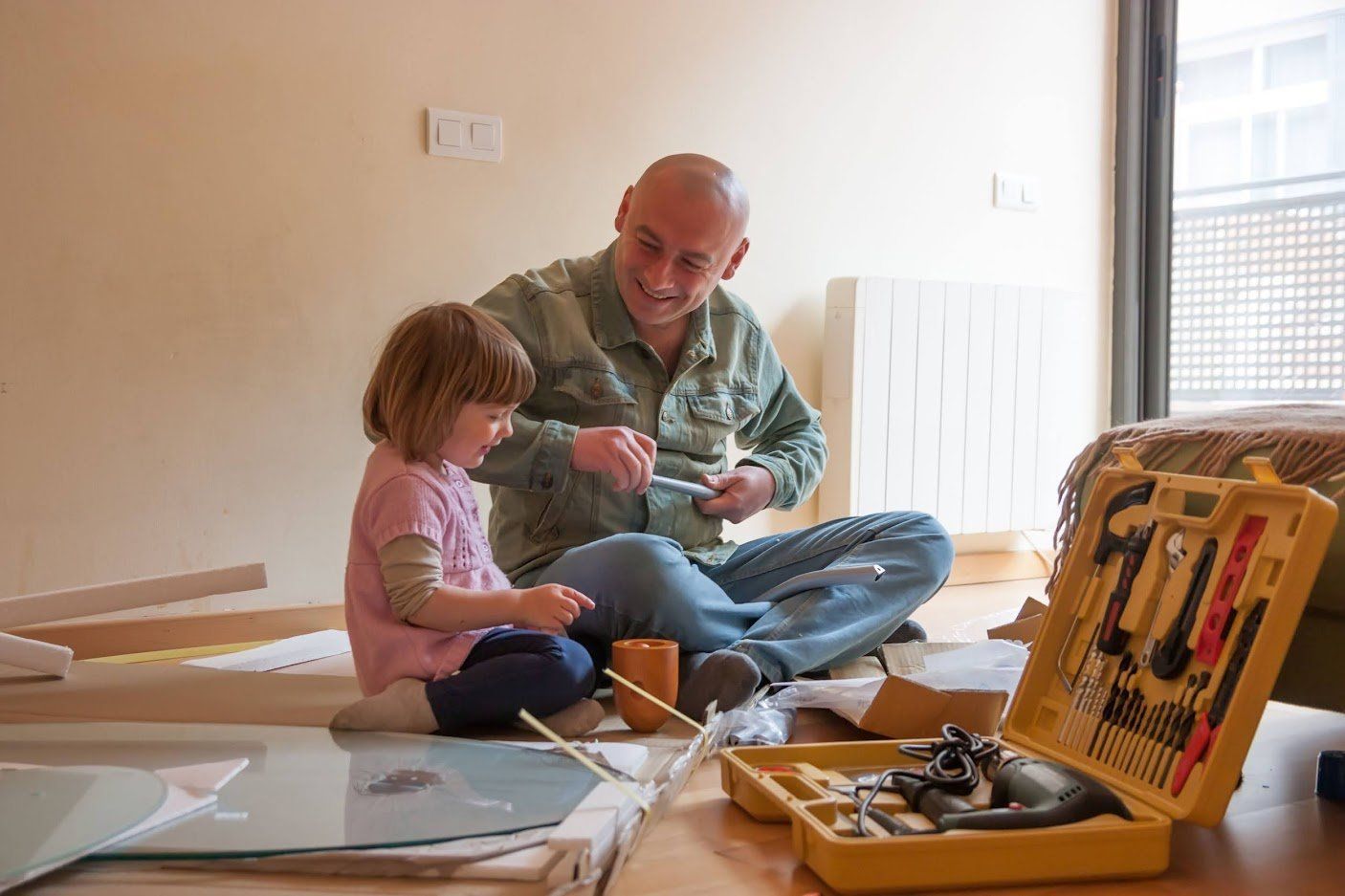 Man and child sitting on the floor, painting together beside open art supplies and a sunlit window.