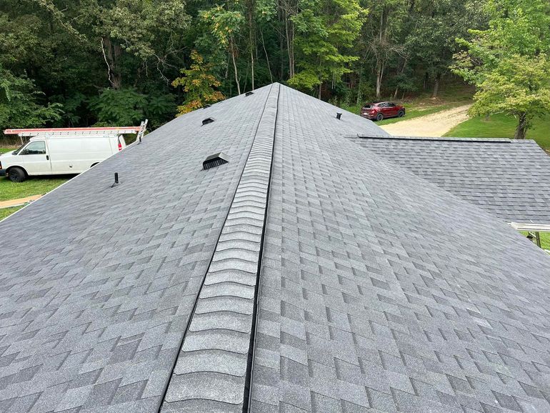 Gray shingled roof with a central ridge vent, viewed from above, with trees and vehicles in the background