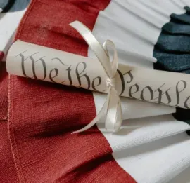Parchment scroll reading “We the People” on red, white, and blue fabric with a rosette ribbon
