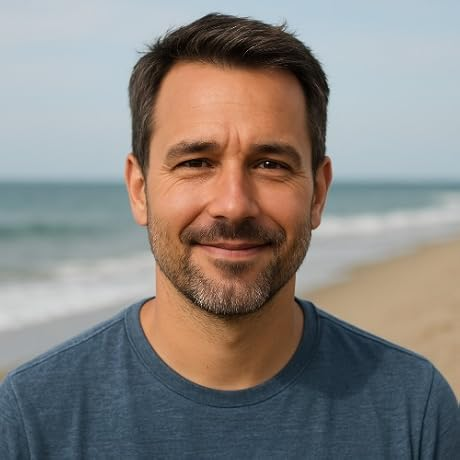 Smiling man in a blue shirt standing on a beach with the ocean behind him