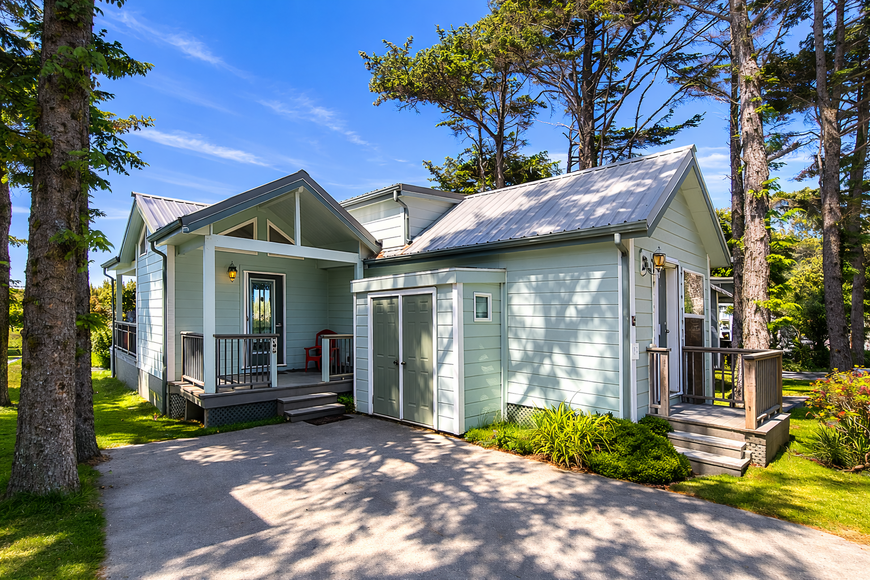Small white cottage with front porch, garage, and trees in a sunny wooded setting
