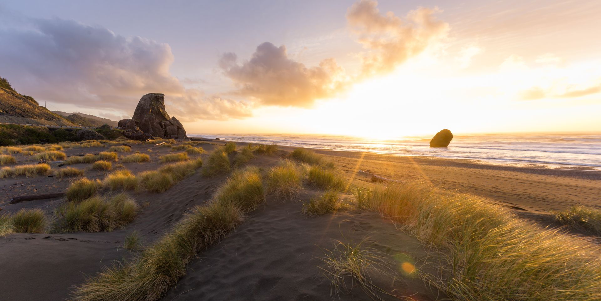 Sunset over a sandy beach with grasses, rocks, and ocean waves glowing in warm light