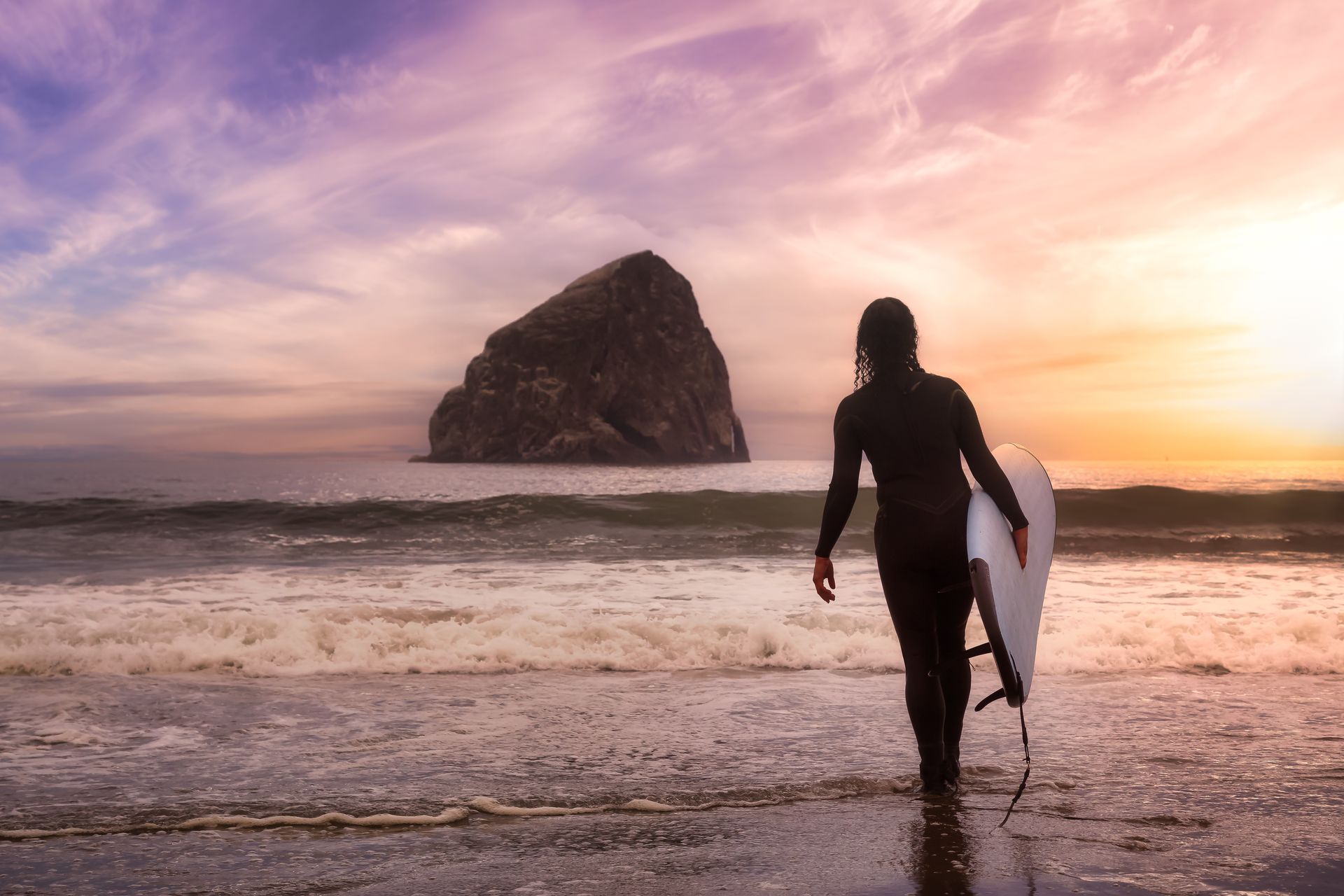 Surfer in a wetsuit walking into the ocean at sunset beside a large sea stack