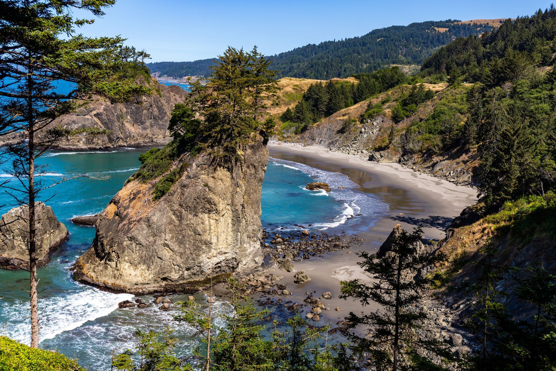 Secluded turquoise cove with sandy beach, rocky cliffs, and evergreen trees under a clear blue sky