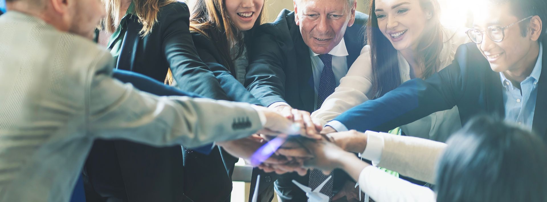 People in business attire stacking hands in a team huddle, smiling in an office setting.
