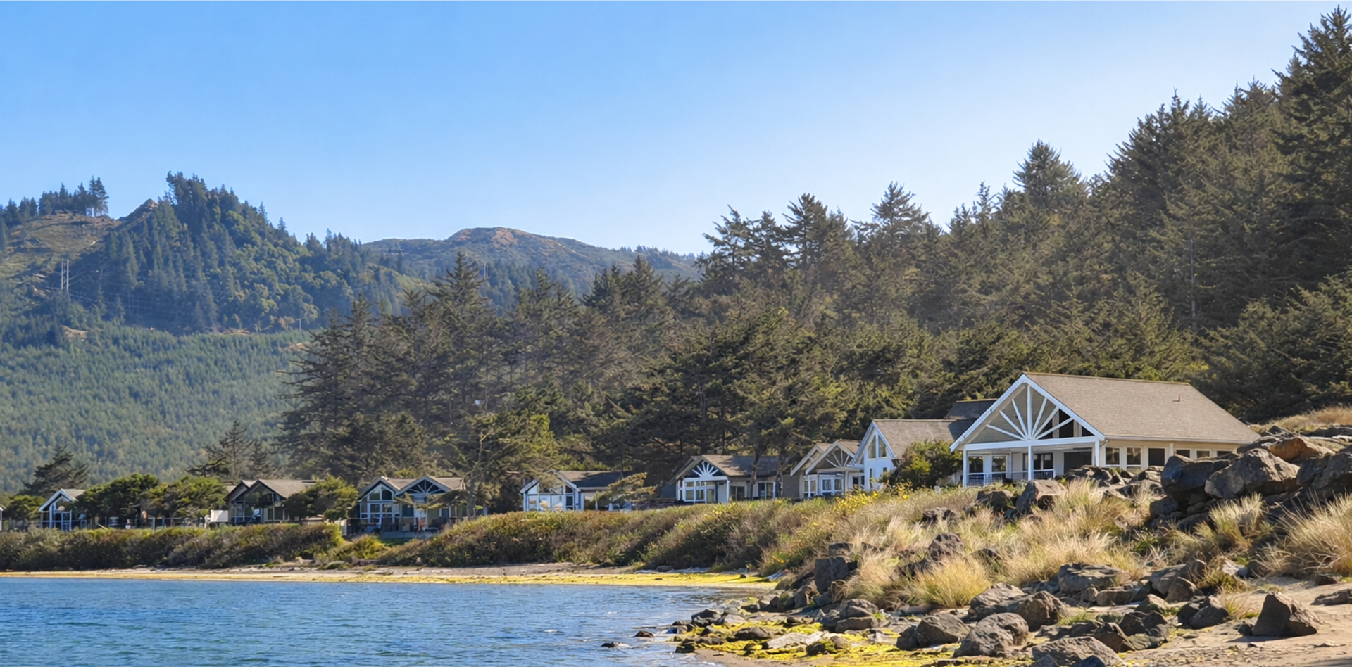 Coastal bay with rocky shore, evergreen trees, and houses at the base of a mountain under a clear sky