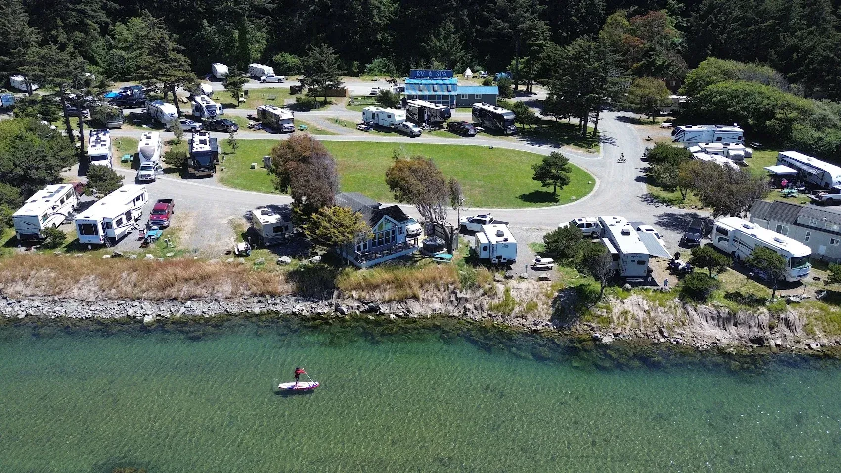 Aerial view of a lakeside campground with RVs, tents, parked cars, and a person canoeing near the shore.