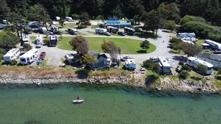 Aerial view of a lakeside campground with RVs, tents, parked cars, and a person canoeing near the shore.