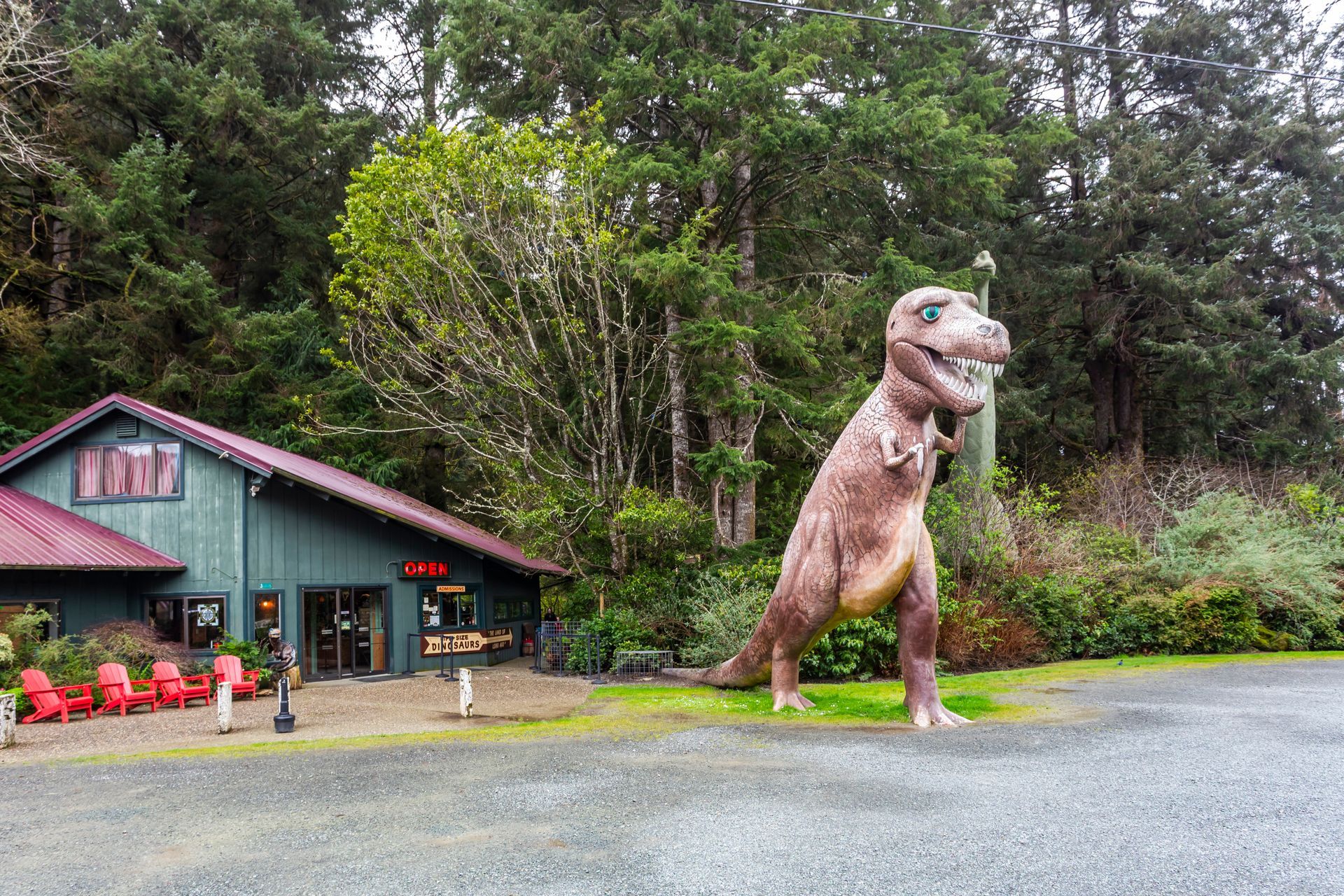 Large dinosaur statue beside a teal building in a forested roadside setting