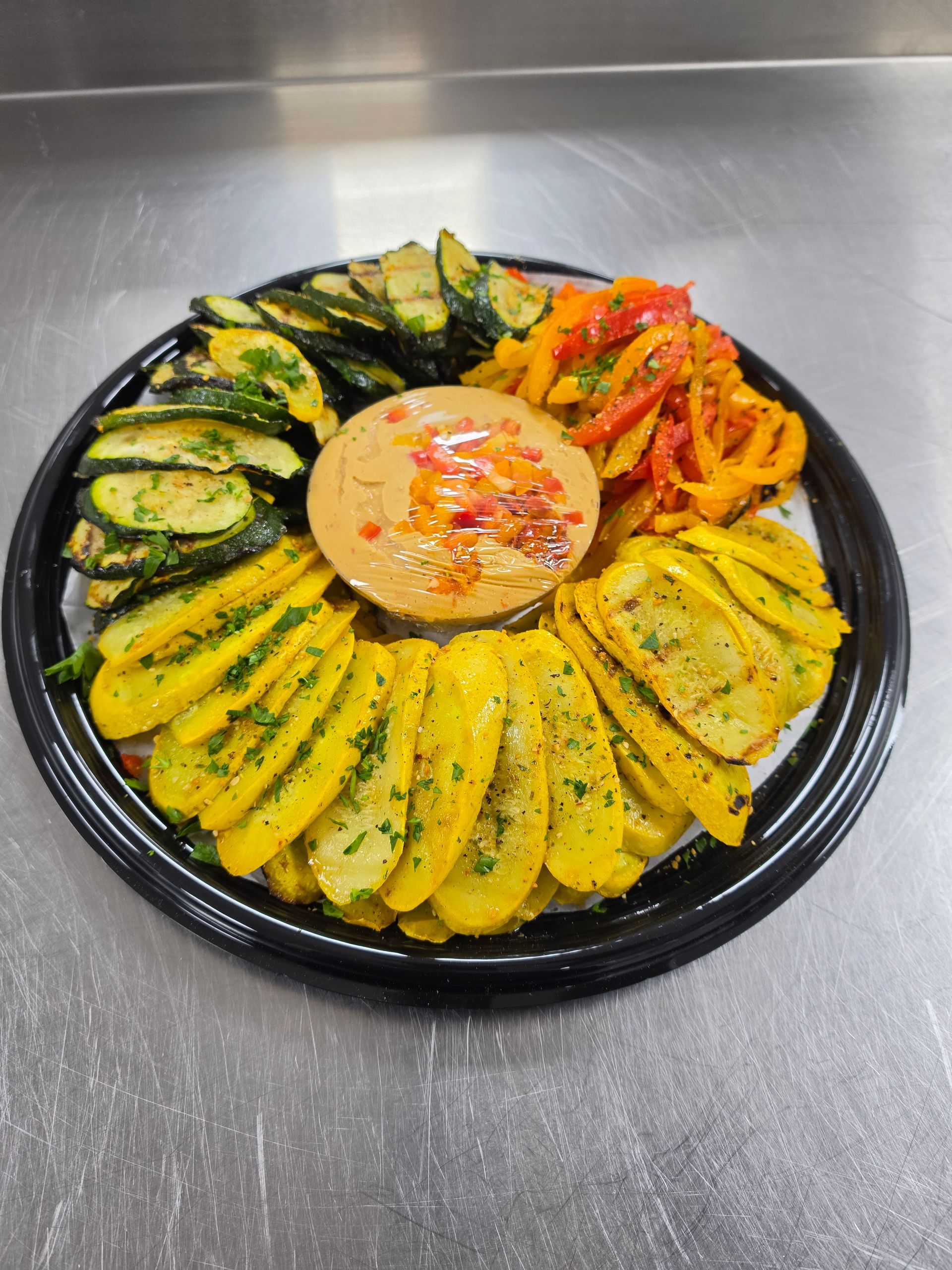 Plated roasted vegetables arranged around a dip on a black tray, with yellow squash, zucchini, and peppers.